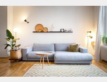 Helles Wohnzimmer mit einer grauen Eckcouch, einem kleinen runden Holztisch und einem cremefarbenen Teppich mit geometrischem Muster. An der Wand befindet sich ein Regal mit dekorativen Körben, Fotos und Trockenblumen, daneben eine Stehlampe und eine Zimmerpflanze im Korb.