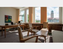 Büro mit großen Fenstern, die viel Tageslicht hereinlassen und den Blick auf ein Gebäude mit Turm in der Ferne freigeben. Der Raum enthält einen Holztisch mit einem Computer, mehrere gepolsterte Stühle, einen niedrigen Couchtisch und ein buntes Gemälde an der Wand.