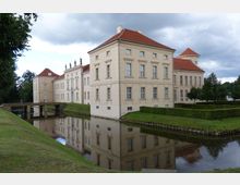Schloss mit cremefarbenen Fassaden und roten Dächern, umgeben von einem Wassergraben, der die Gebäude spiegelt. Im Vordergrund eine Rasenfläche und eine schmale Brücke, die den Graben überquert; im Hintergrund Bäume und ein bewölkter Himmel.