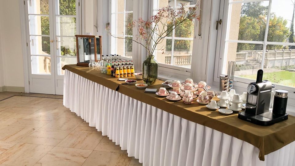 Heller Veranstaltungsraum mit großen, weiß gerahmten Fenstern und Türen, die auf eine Terrasse und einen Parkblick führen. Entlang der Fenster steht ein langer Buffet-Tisch mit Tischdecke, Getränkeflaschen, Porzellan-Tassen und einer Kaffeemaschine.