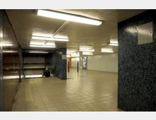Unterirdischer U-Bahn-Bereich mit gefliesten Wänden in Blau- und Weißtönen, einer Treppe, die nach unten führt, sowie einer hellen Beleuchtung durch rechteckige Deckenleuchten. Im Hintergrund ist eine Werbetafel sichtbar.