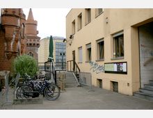 Außenbereich mit einer beige gestrichenen Gebäudefassade, mehreren Fenstern und Graffiti. Links im Bild sind ein roter Backsteinbau mit Türmen, Fahrräder an einem Ständer sowie ein grüner Sonnenschirm zu sehen. Im Hintergrund stehen moderne Gebäude mit Glasfassaden.
