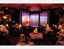 Innenraum eines Cabaret- oder Theatersaals mit runden, weiß eingedeckten Tischen und Stühlen. Im Hintergrund eine Bühne mit glitzerndem Vorhang, darüber eine Leuchtschrift "Kit Kat Klub".