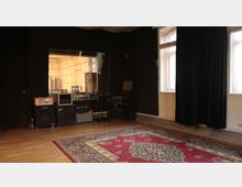 Ein Tonstudio mit schallisolierten Wänden aus Schaumstoff, einem Sichtfenster zum Kontrollraum, mehreren Lautsprechern, Verstärkern und Mikrofonen sowie einem Holzfußboden mit einem großen roten Teppich. Zwei Fenster an der rechten Wand mit Vorhängen lassen Tageslicht hinein.