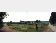 Weite Landschaft mit einem Pferdehof: Im Vordergrund sind Holzlattenzäune, die Koppeln umgeben. In der Mitte steht eine große, flache Stallhalle mit dunkelgrauem Dach. Rechts verläuft ein unbefestigter Weg entlang von Bäumen, im Hintergrund sind Wiesen und Bäume zu sehen.