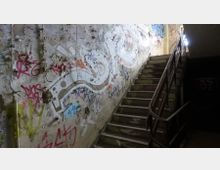 Eine verlassene Treppe in einem heruntergekommenen Gebäude mit abgeplatztem Putz an den Wänden, die mit Graffiti und Zeichnungen übersät sind. Ein Geländer aus Metall führt entlang der Treppe, und Licht fällt von einem oberen Fenster herein.