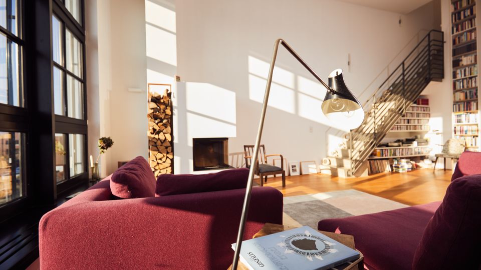 A spacious, modern living room with large floor-to-ceiling windows, burgundy seating, and a minimalist fireplace next to stacked firewood. The room features a staircase leading to a loft area with extensive bookshelves, wooden floors, and mid-century style furniture.
