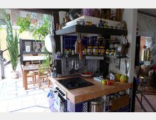 Rustikale Küche mit blau-weißen Fliesen, offenem Regal voller Küchenutensilien und Dekorationen. Im Hintergrund ein Essbereich mit Holztisch, umgeben von Pflanzen und natürlichem Licht.