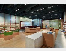 Moderner Apothekeninnenraum mit hellen Holzmöbeln, beleuchteten Regalen voller Produkte und mehreren Bedieninseln mit Touchscreen-Displays. Im Hintergrund befinden sich Glaswände, hinter denen weitere Produkte sichtbar sind.