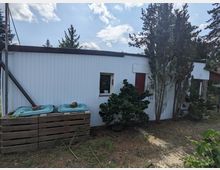 Außenansicht eines niedrigen, weiß verkleideten Nebengebäudes mit Flachdach, kleinem Fenster und roter Tür. Daneben stehen immergrüne Bäume und Sträucher; im Vordergrund sind zwei grüne Abfalltonnen in einer Holzumfassung auf unbefestigtem Boden zu sehen.