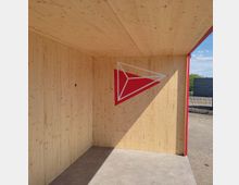 Holzüberdachter Bereich mit hellen Holzpaneelen an den Wänden und der Decke. An der Rückwand ist ein rotes geometrisches Symbol mit weißen Linien angebracht, im Hintergrund ist ein Außenbereich mit Pflastersteinen und Zäunen sichtbar.