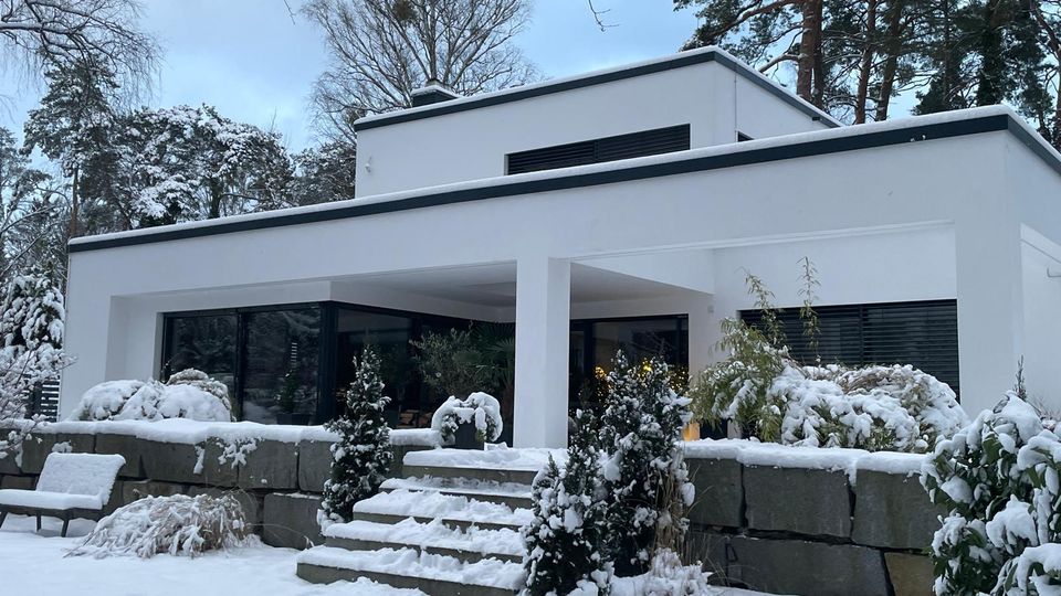 Modernes, weißes Flachdachhaus mit großen Glasfronten und überdachter Terrasse, davor eine niedrige Natursteinmauer mit Treppe. Der verschneite Garten mit Sträuchern und Bäumen umgibt das Gebäude in winterlicher Umgebung.