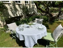 Runder Gartentisch mit weißer Tischdecke und gedecktem Geschirr steht auf einer Rasenfläche neben der Hausfassade mit Fenstern und einem Blumenbeet. Im Hintergrund sind ein großer Baum, eine niedrige Natursteinmauer und weitere Gartenpflanzen zu sehen.
