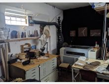 Ein kleiner Werkstattraum mit einer Werkbank und einer gravierten Laser-Schneidemaschine. Im Vordergrund sind Werkzeuge, Holzstücke und Materialien verstreut, während an den Wänden Regale mit Gegenständen und eine kleine Fensteröffnung mit Gitter zu sehen sind.