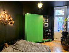 Schlafzimmer mit dunklen Wänden, einem Bett mit gestreifter Bettwäsche und einem grünen Kleiderschrank. Rechts befindet sich ein Fenster mit Blick nach draußen, daneben stehen Pflanzen und ein Regal mit Schuhen auf einem Holzboden.