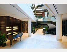 Eingangsbereich eines modernen Gebäudes mit einem schwarzen Flügel, geschwungener Treppe mit Glasgeländern und Marmorböden. Die Wände sind teilweise mit dunklem Holz und Marmor verkleidet, dekoriert mit Pflanzen und einer Beschilderung.