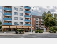 Wohnhaus mit mehreren Stockwerken, weißen Fassaden, blauen Balkonen und einem Café im Erdgeschoss mit gelben Markisen. Im Hintergrund steht ein weiteres Wohngebäude, umgeben von Bäumen und Grünflächen, daneben eine ruhige Straße.