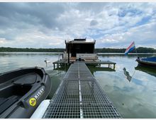 Ein Steg führt zu einem modernen Hausboot auf einem See, umgeben von bewaldetem Ufer. Im Vordergrund ist ein schwarzes Ruderboot zu sehen, während auf der rechten Seite eine niederländische Flagge an einem weiteren Boot weht.
