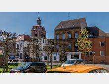 Straßenszene mit historischen Gebäuden, darunter ein roter Kirchturm mit einer Turmspitze und eine Fassade aus rotem Ziegelstein. Im Vordergrund geparkte Autos, Laternen und geschnittene Bäume entlang eines Gehwegs.