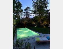 Ein moderner Garten mit einem rechteckigen, beleuchteten Swimmingpool bei Dämmerung, umgeben von Bäumen und Rasenflächen. Im Vordergrund steht eine Liege mit farbigen Kissen auf einer gepflasterten Terrasse.