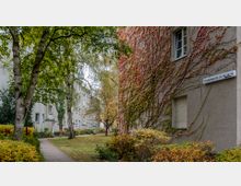 Wohnsiedlung mit mehreren hellen, mehrstöckigen Gebäuden, umgeben von Bäumen und gepflegten Grünflächen. An einer Hauswand wachsen Kletterpflanzen, die herbstlich verfärbt sind, und ein Straßenschild mit der Adresse "Schöppinger Str. 20–14" ist sichtbar.