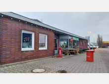 Ein einstöckiges Backsteingebäude mit Flachdach, großen Fenstern und einem Eingang mit Glastür. Vor dem Gebäude stehen Sitzgelegenheiten, Blumentöpfe sowie rote Fass-Tische, und der gepflasterte Vorplatz grenzt an einen Parkplatz.