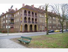 Wohngebäude aus der frühen Moderne mit brauner Fassade, gelben Fensterrahmen und dekorativen Balkonen mit Bögen und gemusterten Geländern. Im Vordergrund befinden sich eine Parkbank, eine Straße und geparkte Autos, umgeben von kahlen Bäumen und Grünflächen.