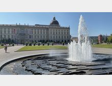 Brunnen mit sprudelndem Wasser im Vordergrund, dahinter ein historisches Gebäude mit einer großen Kuppel und kunstvoller Fassade, umgeben von einer Rasenfläche und Spazierwegen. Der blaue Himmel deutet auf sonniges Wetter hin.