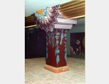 Eine dekorative Stütze in einem Innenraum, gestaltet wie ein stilisierter Baumstamm mit roten und blauen Mustern, sowie einem kunstvoll verzierten Kapitell in Form eines Mosaiks aus bunten Blättern und einem Sternmotiv. Der Boden besteht aus beigefarbenen Fliesen, und im Hintergrund sind Wandmosaike mit pflanzlichen und geometrischen Motiven zu sehen.