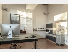 Modernes Büro mit minimalistischer Einrichtung, darunter ein großer Schreibtisch mit einem Desktop-Computer, ein Schränkchen mit Schubladen und eine Kaffeemaschine. Die Wände sind weiß, mit gerahmten Kunstwerken und Fotos dekoriert, und natürliches Licht fällt durch große Fenster ein.