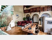 Innenraum eines Wohnbereichs in einem historischen Gebäude mit sichtbarer Holzvertäfelung und dekorativen Bögen. Im Vordergrund steht ein großer Holztisch mit Stühlen, im Hintergrund befinden sich ein rotes Sofa, Bücherregale und eine weiße Tür unter Rundbögen.