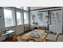 Ein einfacher Raum mit großen Fenstern, die viel Tageslicht hereinlassen, und einer weißen Wand, an der alte landwirtschaftliche Werkzeuge hängen. Ein Tisch mit verstreuten Papieren und mehreren Stühlen sowie eine an der Wand stehende Leiter prägen die Einrichtung.