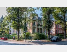 Straßenszene mit einem historischen Mehrfamilienhaus im Stil der Gründerzeit, gekennzeichnet durch helle Stuckverzierungen und rotes Mauerwerk. Umgeben von Bäumen und Grünflächen, stehen geparkte Autos am Straßenrand, und Passanten gehen auf dem Gehweg spazieren.