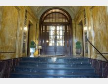 Eingangsbereich eines historischen Gebäudes mit einer Treppe aus dunklem Marmor, Wänden aus gelblichem poliertem Stein und einer großen Holztür mit Glas- und Metallrahmen. Zwei dekorative Pflanzen in Töpfen flankieren die Tür, und eine Teppichläufer liegt auf dem Boden.