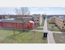 Luftaufnahme eines Campusgeländes mit einer Mischung aus roten Backsteingebäuden und modernen gelb-beigen Gebäuden. Im Vordergrund sind Grünflächen, vereinzelte Bäume und gepflasterte Gehwege zu sehen, die das Gelände durchziehen.