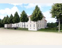 Großer, heller Schlossbau im klassischen Stil mit weißen Fassaden und roten Dächern, umgeben von einer gepflegten Gartenanlage mit symmetrisch angeordneten Bäumen und Hecken. Im Vordergrund ist ein Kiesweg und eine kleine Brücke über einen Wassergraben zu sehen.