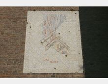 Mosaikrelief an einer Ziegelmauer, das ein stilisiertes, brennendes Feuer mit Flammen in roten und grauen Tönen zeigt. Unterhalb des Bildes steht in roten Buchstaben „DAS FEUER“.