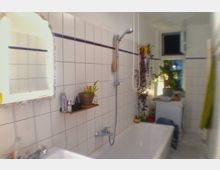 Ein schlichtes Badezimmer mit weiß gefliesten Wänden, einer Badewanne mit Duschkopf und einem Spiegel an der Wand. Pflanzen und Pflegeprodukte befinden sich auf einem kleinen Regal und an der Fensterbank; das Fenster ist leicht geöffnet.