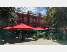 Historisches Backsteingebäude mit verzierten Fenstern und gläsernen Dachstrukturen im Hintergrund, davor ein Platz mit roten Sonnenschirmen, Sitzplätzen, Fahrrädern und umgebender Begrünung.