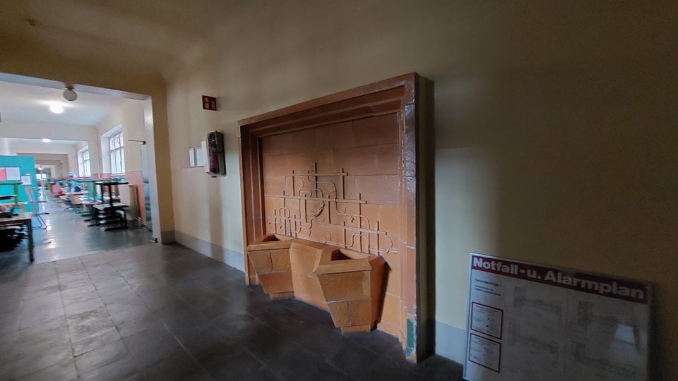 Ein Flur mit einem eingebauten Brunnen aus orangefarbenen Fliesen, verziert mit geometrischen Mustern. Im Hintergrund sind helle Fenster, Tische und Stühle eines Arbeitsbereichs zu sehen; an der Wand hängt ein Feuerlöscher sowie ein Notfall- und Alarmplan.