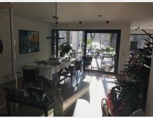 Moderner Essbereich in einem Wohnhaus mit großem Esstisch und dunklen Stühlen, daneben bodentiefen Schiebetüren zur sonnigen Terrasse mit Pflanzen. Rechts steht ein dekorierter Weihnachtsbaum; große Fensterflächen lassen viel Licht in den Raum.