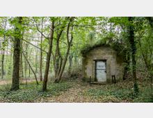 Ein kleines, rechteckiges Gebäude aus Stein mit der Inschrift „Römisches Wasserwerk“ auf der Fassade, umgeben von dichtem Wald mit Laubbäumen und Bodendeckerpflanzen. Die Umgebung wirkt überwachsen, und das Gebäude scheint verlassen.