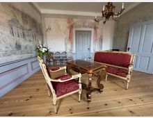 Barockstil-Zimmer mit Holzboden, einer gold-verzierten Sitzgarnitur mit rotem Stoffbezug und einem aufwendigen Holztisch. Die Wände sind mit historischen Wandmalereien dekoriert, und ein Kronleuchter hängt an der Decke.