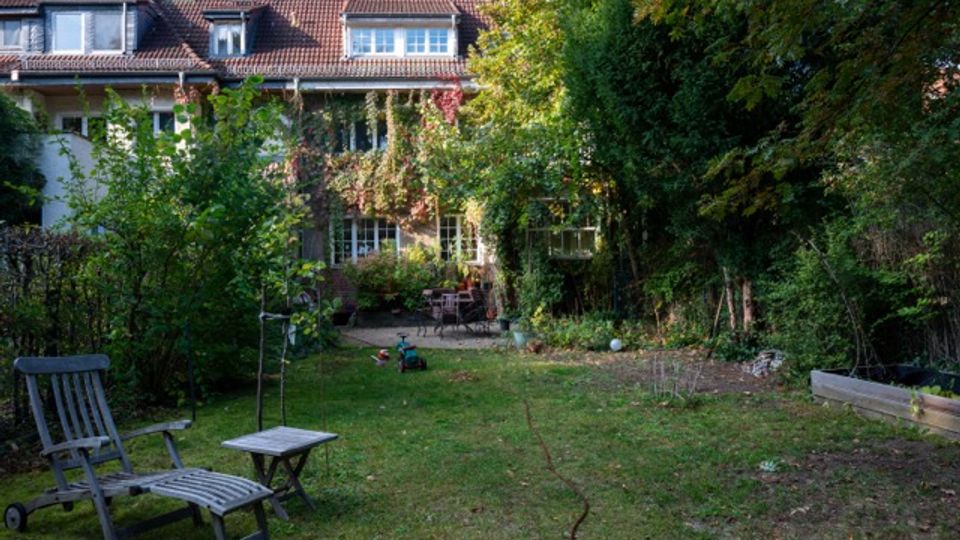 Großer Garten hinter einem mehrstöckigen Wohnhaus mit rotem Ziegeldach und vielen Fenstern, teils von Kletterpflanzen bewachsen. Auf der Rasenfläche stehen Gartenliegen und ein kleiner Tisch; dichte Bäume und Sträucher säumen die Grundstücksgrenzen.