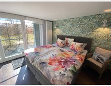 Ein Schlafzimmer mit einem Doppelbett, das bunte florale Bettwäsche hat, und einer grünen Tapete mit Pflanzenmuster an der Wand. Große Fenster lassen Tageslicht herein und bieten einen Blick auf eine Hecke und eine Terrasse; daneben steht ein Korbstuhl mit einem Kissenmotiv.