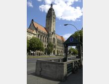 Ansicht eines historischen Gebäudes mit einem hohen Turm und rotem Ziegeldach, im Vordergrund der Eingang zur U-Bahn-Station "Richard-Wagner-Platz" mit einer grauen Steinbrüstung und Fahrrädern. Die Straße, Bäume und einige parkende Autos umgeben die Szene.