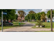 Eine Kopfsteinpflasterstraße führt durch eine Dorfidylle mit gepflegten Grünflächen, üppigen Bäumen und historischen Gebäuden im Hintergrund, darunter ein großes, rosafarbenes Haus. Ein Straßenschild mit der Aufschrift „An der Dorfau“ ist sichtbar, flankiert von geparkten Autos und Blumenkübeln.