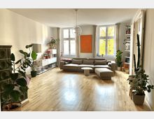 Geräumiges Wohnzimmer mit hellem Holzboden, einer großen grauen Eckcouch und bodentiefen Fenstern mit blickdichten Vorhängen. Es gibt mehrere grüne Zimmerpflanzen, einen grünen Kachelofen, ein Wandregal und moderne Dekorationen, darunter ein auffälliges orangefarbenes Kunstwerk.