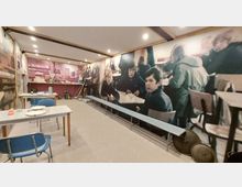 Innenraum einer Ausstellung, die eine historische Schulspeisung darstellt, mit Tischen, einfachen Metallstühlen und Geschirr auf den Tischen. Die Wände sind mit großformatigen Fotografien von Schülern in einem Speisesaal dekoriert, im Hintergrund steht eine Nachbildung eines Lebensmittelverkaufsbereichs mit Waage und Utensilien.