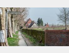 Ein schmaler Gehweg führt entlang einer Baumreihe mit kahlen Ästen und liegt neben einer Wiese, die von einem Holzzaun abgegrenzt ist. Im Hintergrund sind mehrere Backsteinhäuser und ein Fachwerkhaus zu sehen, ergänzt durch einen Straßenname-Schild mit der Aufschrift "Hellriegelstraße".